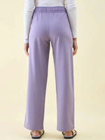 Model wears 98degreenorth Violet Wide Leg Pintuck Trackpants, white long-sleeve top, and sandals, shown from the back.