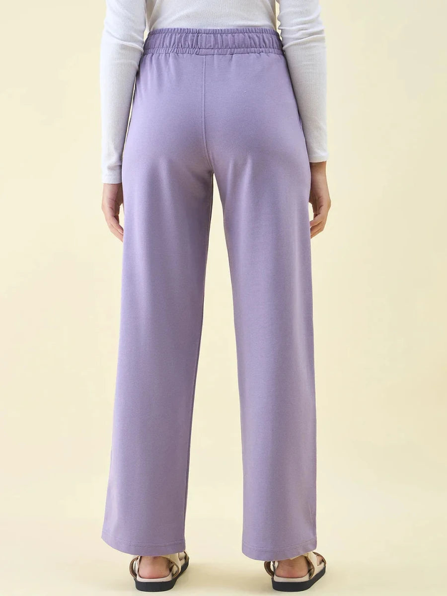 Model wears 98degreenorth Violet Wide Leg Pintuck Trackpants, white long-sleeve top, and sandals, shown from the back.