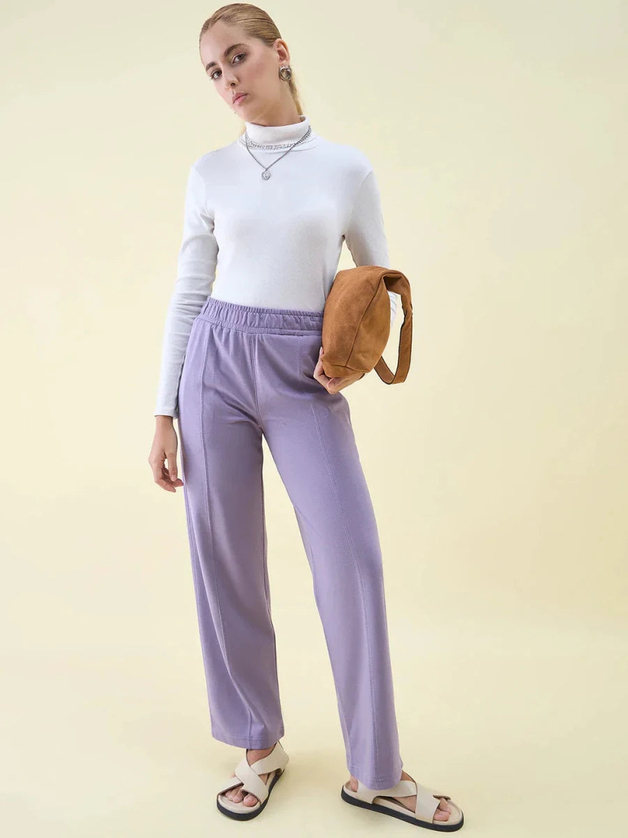 Woman in a white turtleneck and 98degreenorth Violet Wide Leg Pintuck Trackpants, white sandals, holding a brown bag.