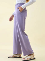 Person in 98degreenorth Violet Wide Leg Pintuck Trackpants, Luxe Lounge style, white top, beige sandals, hand in pocket.