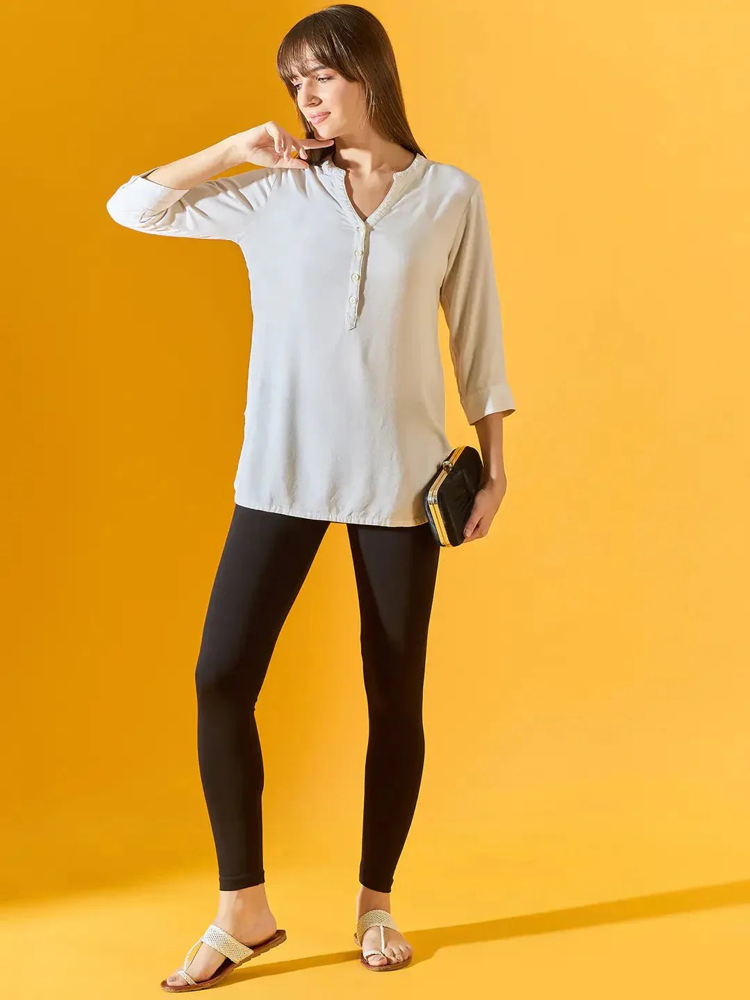 A woman stands against a yellow background wearing 98degreenorth Women Black Ankle-Length Leggings, a light beige tunic, white sandals, and holding a small black clutch. She gazes to the side, one hand touching her hair.
