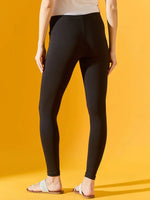 A person stands against a yellow background wearing 98degreenorth Women Black Ankle-Length Leggings, a white top, and white open-toe sandals. The back view highlights the sleek, stretchable fit of these leggings for women.