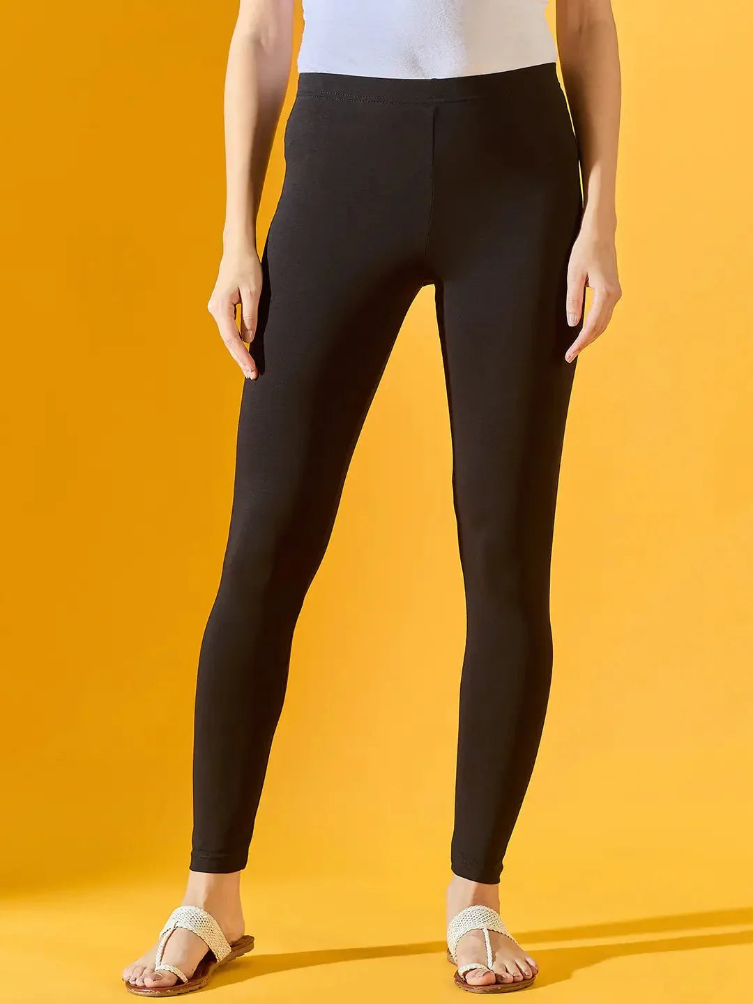 A person in a white top and sandals stands against a yellow background, showcasing the lower half dressed in 98degreenorth Women Black Ankle-Length Leggings.