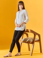 A woman with straight brown hair stands by a wooden chair, wearing 98degreenorth Women Black Ankle-Length Leggings, a light long-sleeve tunic, and white sandals against a yellow background.
