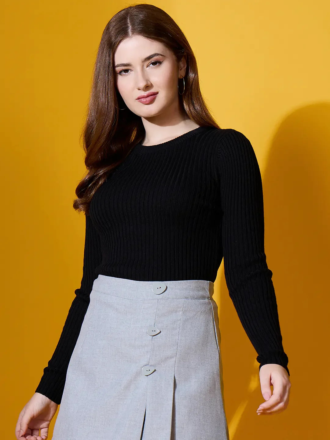 Woman in a 98degreenorth black ribbed round neck sweater and light gray skirt stands by a yellow background.