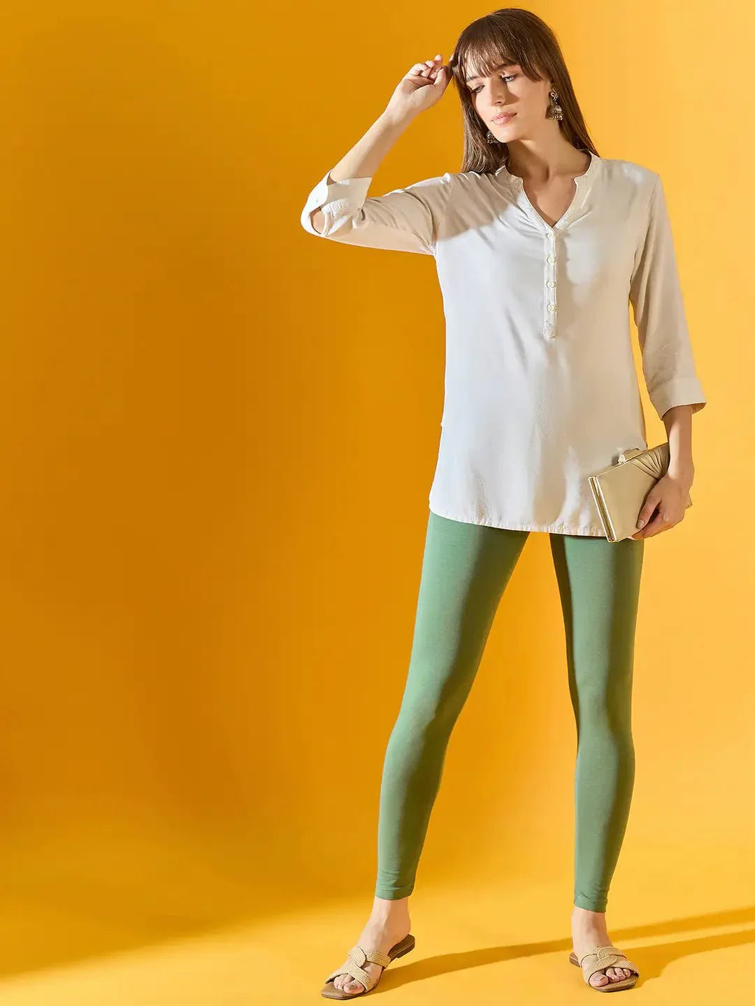 Woman in cream tunic and 98degreenorth Women Bright Green Ankle-Length Leggings holds beige clutch against yellow backdrop.