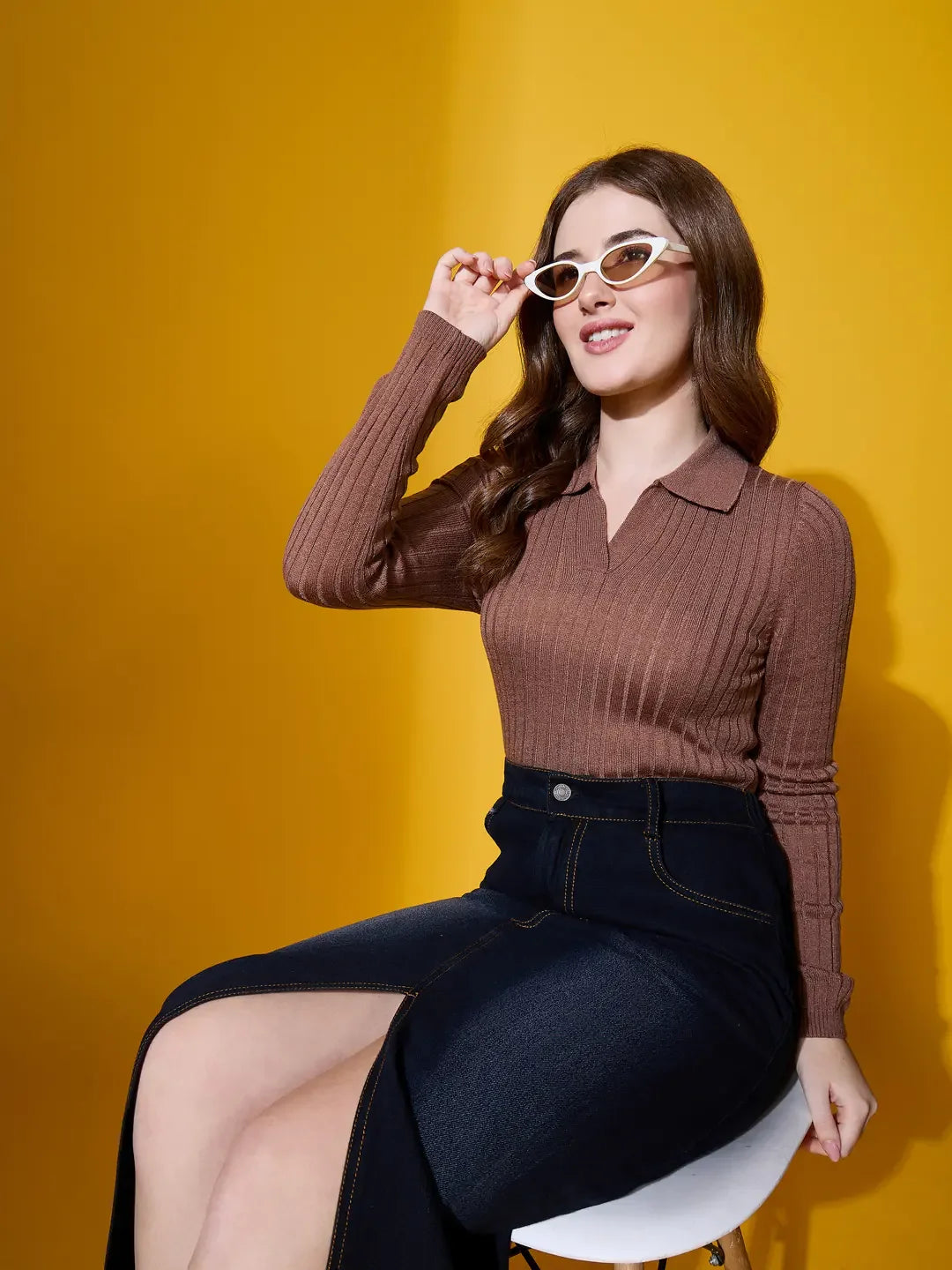 Woman in 98degreenorth camel ribbed collar sweater and dark skirt adjusts white sunglasses against a yellow background.