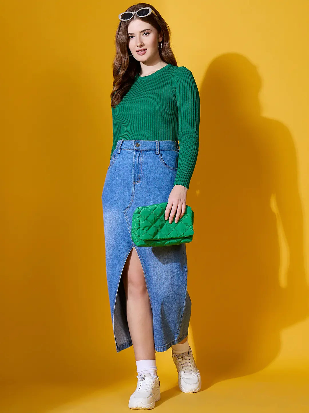 Woman in a 98degreenorth emerald green ribbed sweater and skirt, holding a clutch, stands by a yellow wall.