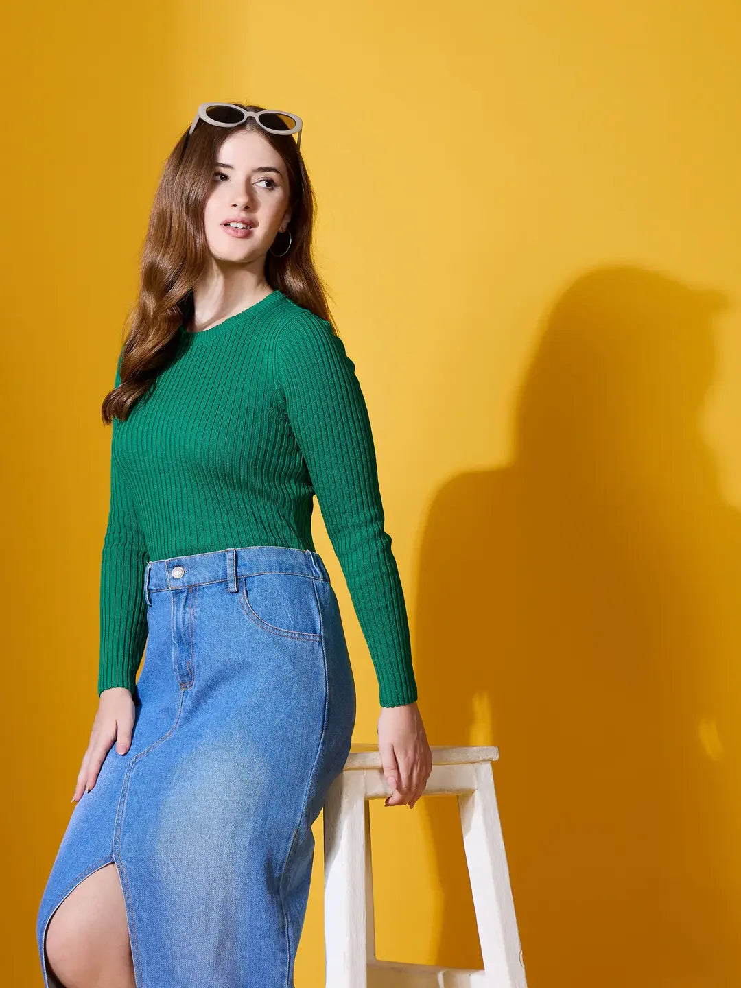 Woman wears 98degreenorth Emerald Green Ribbed Sweater, sitting on a stool against a yellow background, sunglasses atop her head.