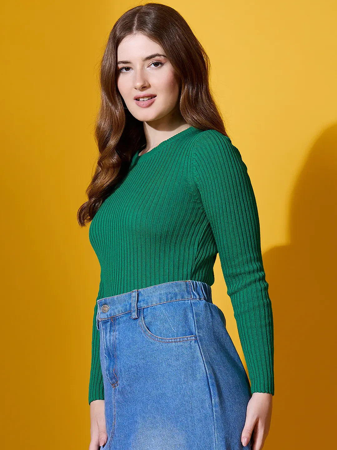 A woman in the 98degreenorth Emerald Green Ribbed Sweater and blue jeans stands before a yellow background.