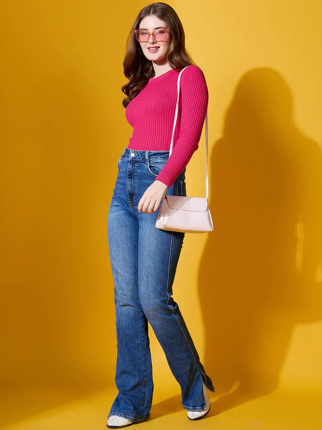 Woman smiles in a 98degreenorth fuchsia ribbed sweater, blue jeans, and glasses with a white purse against yellow.
