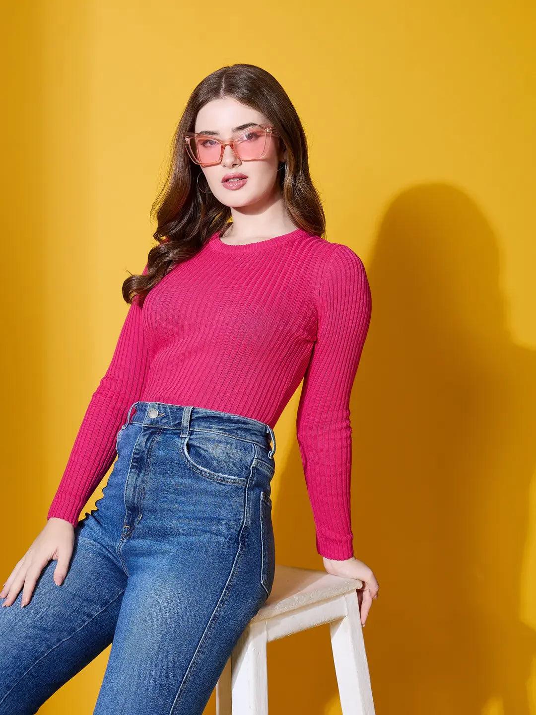 Woman in fuchsia 98degreenorth ribbed sweater and jeans sits on a stool, pink sunglasses, yellow background.