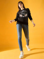 A woman poses against a yellow background, wearing a 98degreenorth Women Graphic Embroidered Black Round Neck Fleece Sweatshirt, blue skinny jeans, and white sneakers.