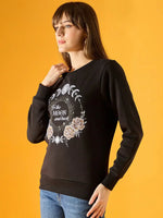 A woman with straight brown hair and blue jeans wears a 98degreenorth Women Graphic Embroidered Black Round Neck Fleece Sweatshirt featuring a moon phase and floral design with the text To the MOON and back, standing against a yellow background.