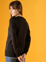 A woman with straight brown hair wears the 98degreenorth Women Graphic Embroidered Black Round Neck Fleece Sweatshirt and blue jeans, standing against a yellow background and looking over her shoulder with her hands behind her back.