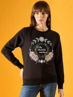 A woman wears the 98degreenorth Women Graphic Embroidered Black Round Neck Fleece Sweatshirt, featuring celestial and floral designs with “To the moon and back” on the front, against a yellow background.