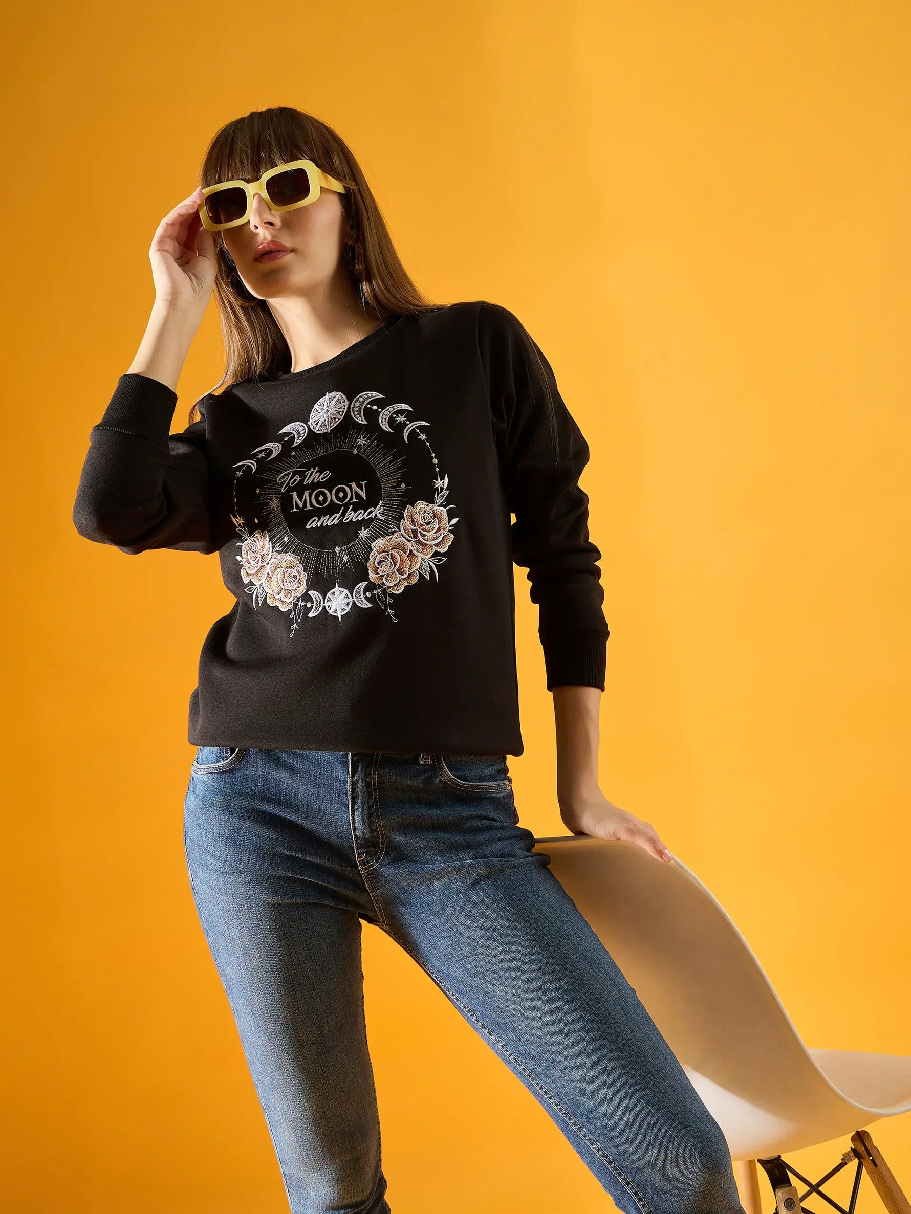 A woman in sunglasses poses confidently against a yellow background, holding a white chair while wearing the 98degreenorth Women Graphic Embroidered Black Round Neck Fleece Sweatshirt.