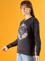 A woman with straight brown hair wears a 98degreenorth Women Graphic Embroidered Navy Round Neck Fleece Sweatshirt with moon phases, roses, and To the moon and back text, paired with blue jeans against a bright yellow background.