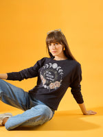 A woman with long brown hair sits on the floor against a yellow background, wearing a 98degreenorth Women Graphic Embroidered Navy Round Neck Fleece Sweatshirt, light blue jeans, white sneakers, and sunglasses on her head.