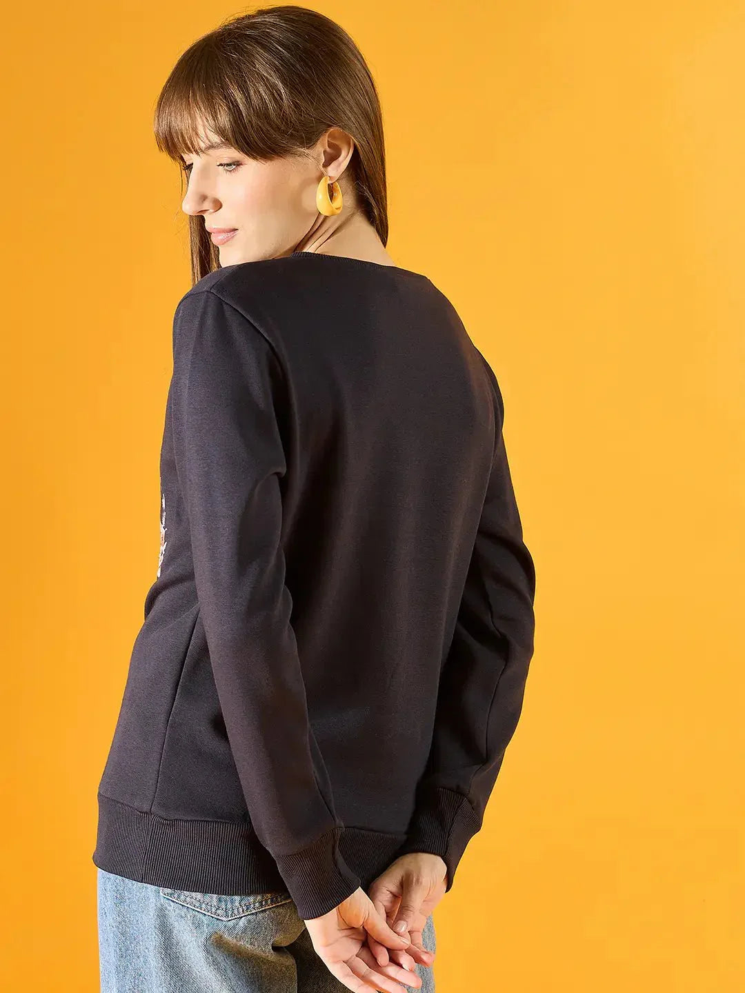 A woman with straight brown hair and yellow earrings poses against a yellow background, wearing the 98degreenorth Women Graphic Embroidered Navy Round Neck Fleece Sweatshirt with blue jeans.