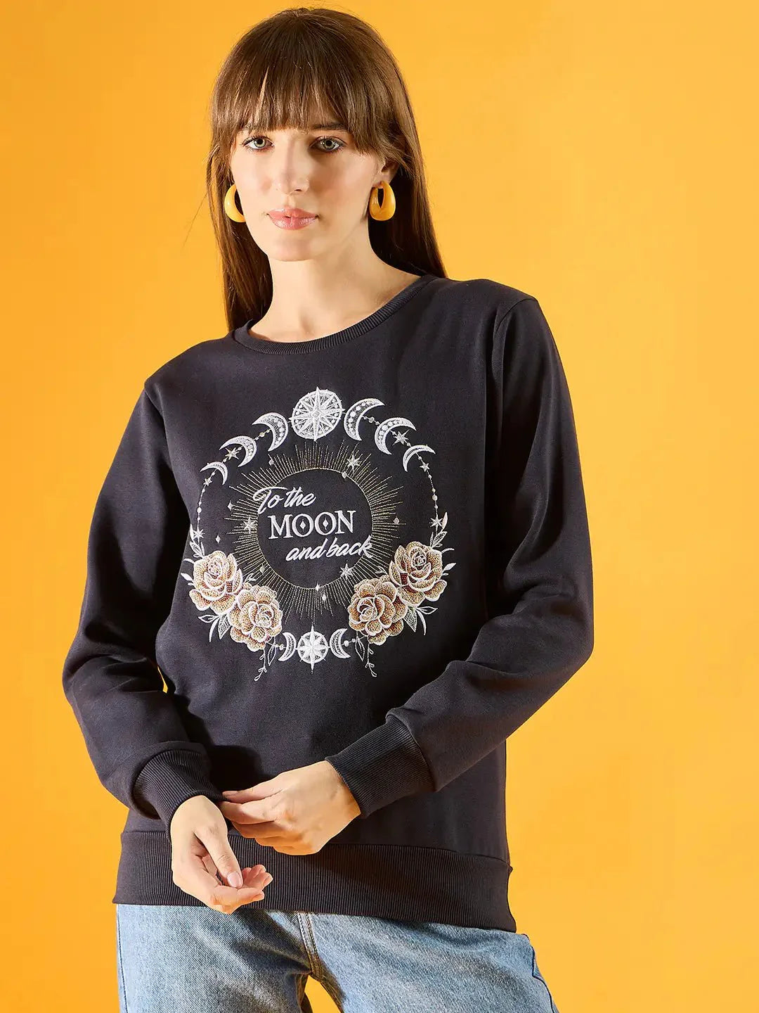 A woman with straight brown hair wears yellow hoop earrings and a 98degreenorth Women Graphic Embroidered Navy Round Neck Fleece Sweatshirt, featuring “To the Moon and back” with moon phases and roses, against a solid yellow background.