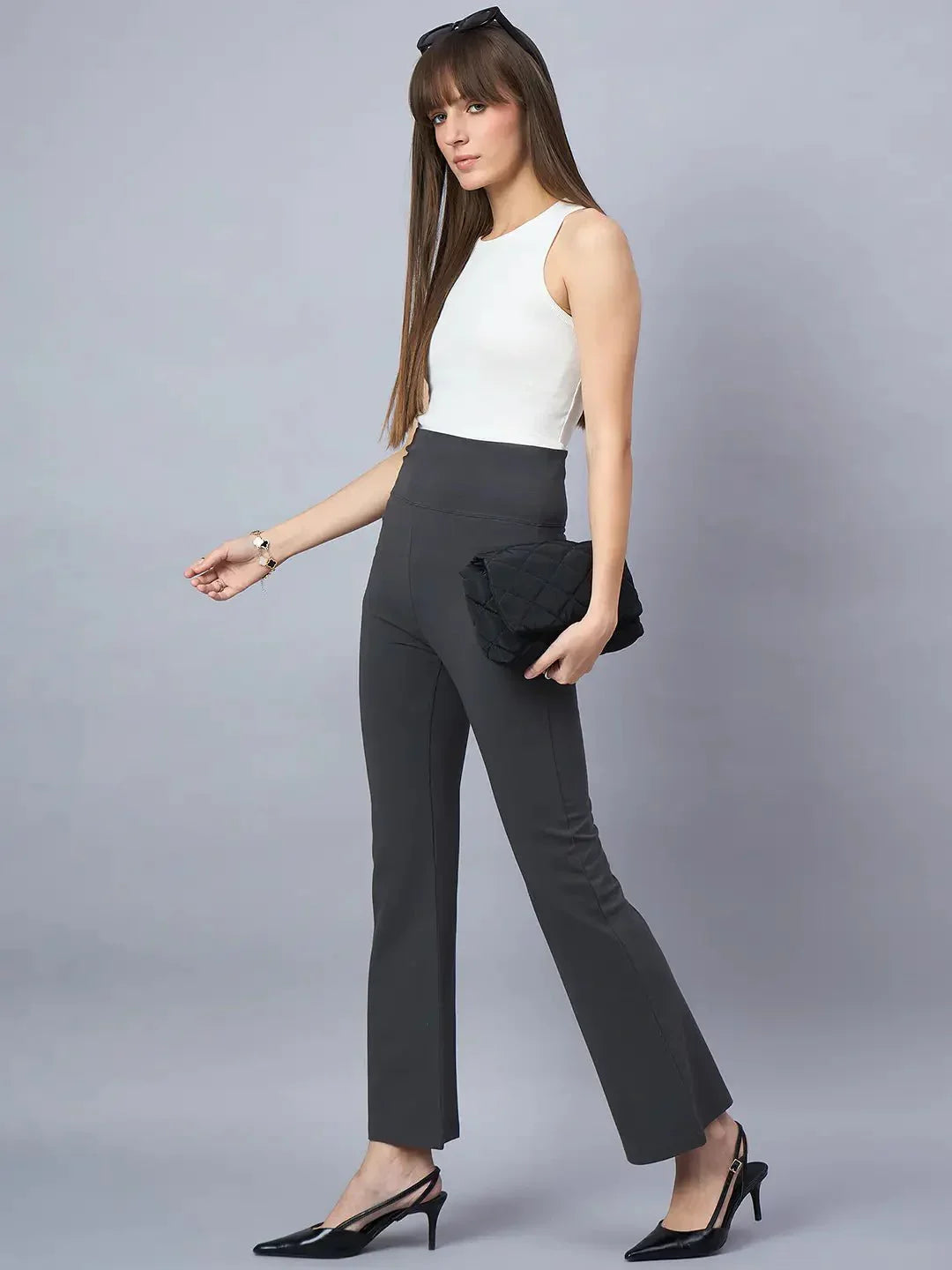 A woman with long straight brown hair and bangs stands against a gray background wearing a sleeveless white top, 98degreenorth Women High-Rise Charcoal Cotton Trousers, black heels, and holding a black clutch.