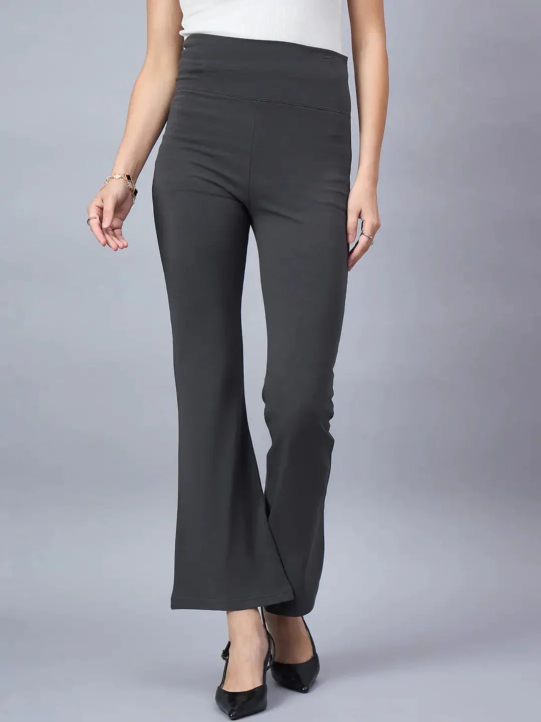 A woman stands against a light grey background wearing 98degreenorth Women High-Rise Charcoal Cotton Trousers, black pointed-toe heels, and a white top.