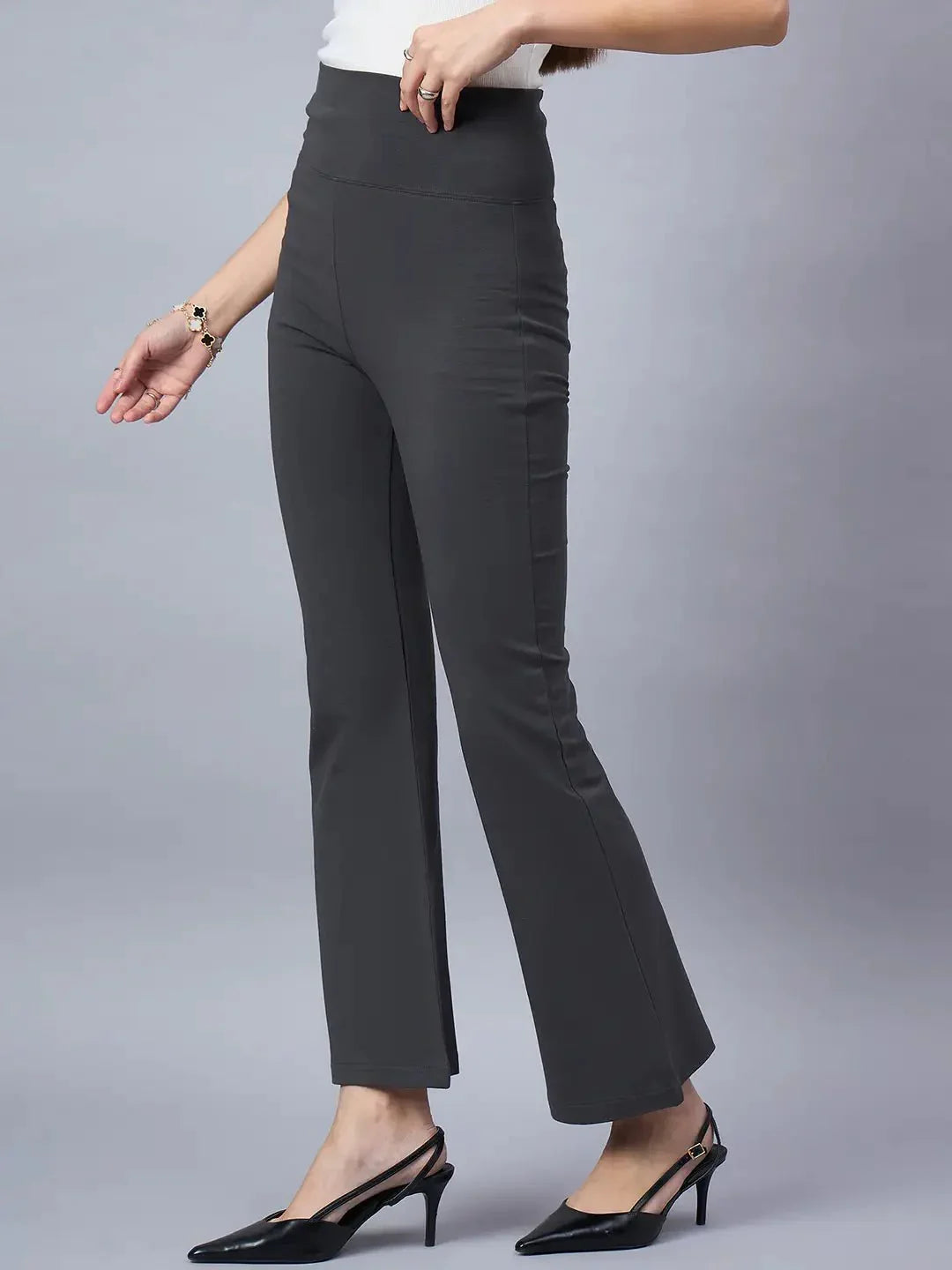 A woman models 98degreenorth Women High-Rise Charcoal Cotton Trousers, styled with a white top, black heels, and a bracelet, set against a plain gray background.
