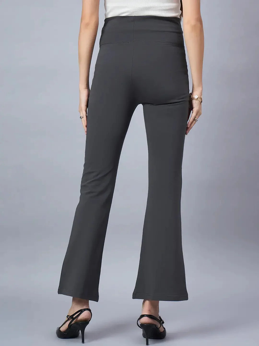 A woman faces away, showcasing 98degreenorth’s Women High-Rise Charcoal Cotton Trousers with a flared silhouette and black heels, paired with a sleeveless light top against a plain gray background.