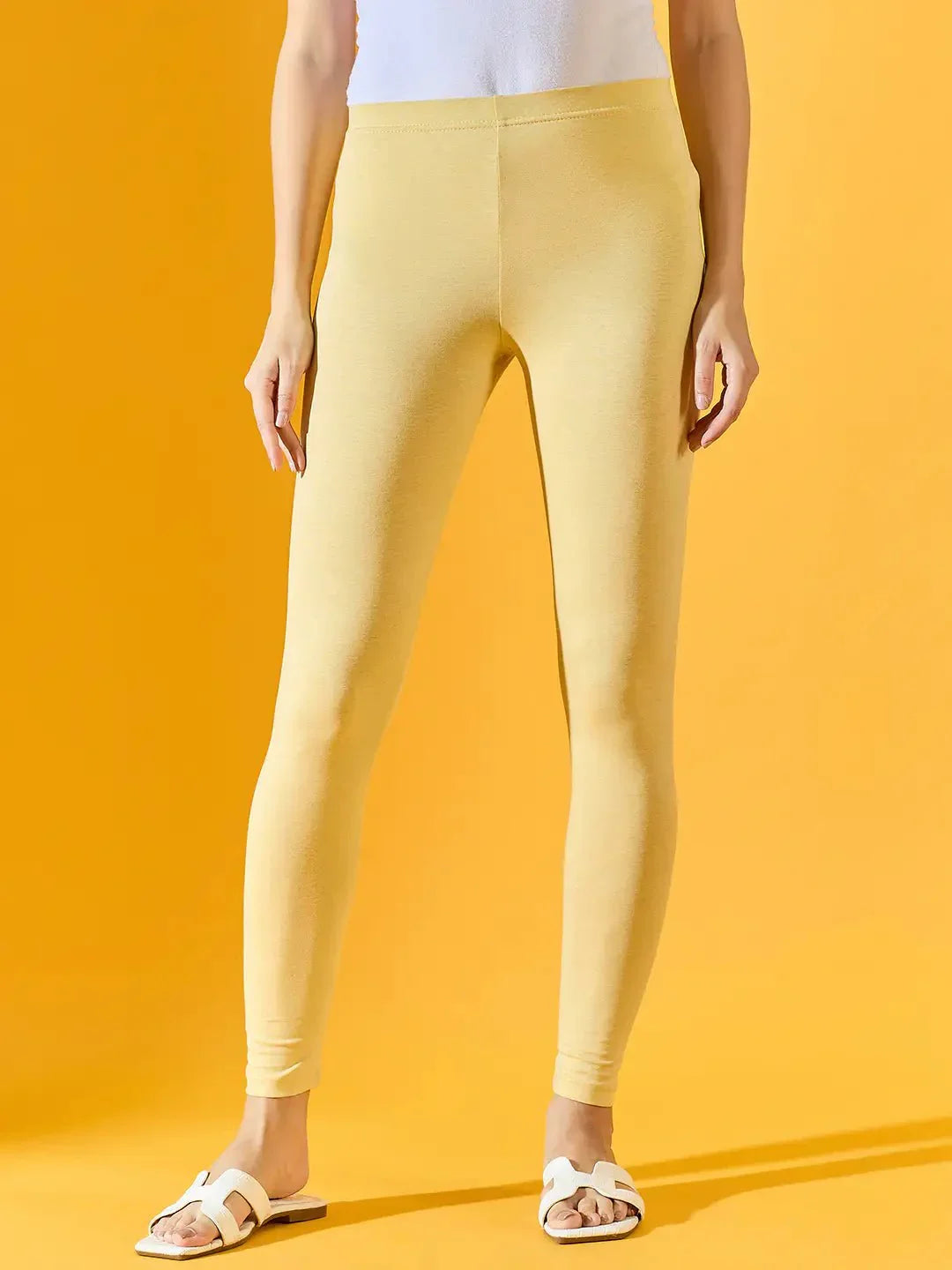 A person in 98degreenorth Women Mango Ankle-Length Leggings, a white top, and sandals stands by a yellow background.