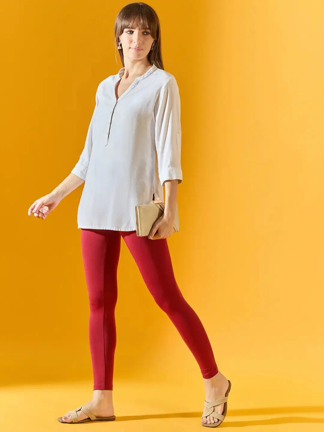 Woman in maroon ankle-length leggings by 98degreenorth holds a beige clutch against a yellow background.