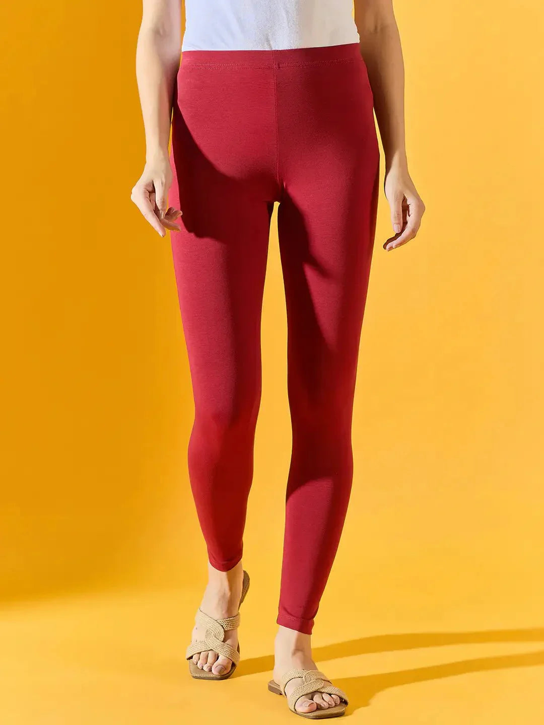 Person wearing 98degreenorth Women Maroon Ankle-Length Leggings, a white top, and beige sandals against a yellow background.