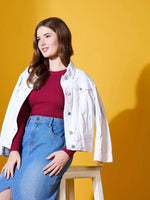 Woman in a 98degreenorth maroon round neck ribbed sweater and blue denim skirt sits on a stool against yellow.