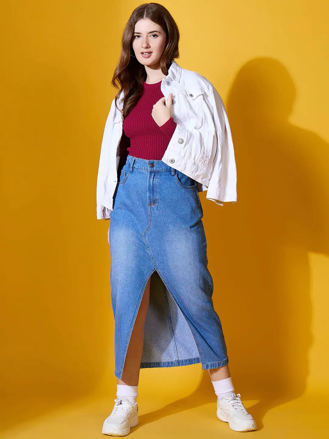 A woman wears the 98degreenorth maroon ribbed sweater, styled with a denim skirt and white sneakers against yellow.