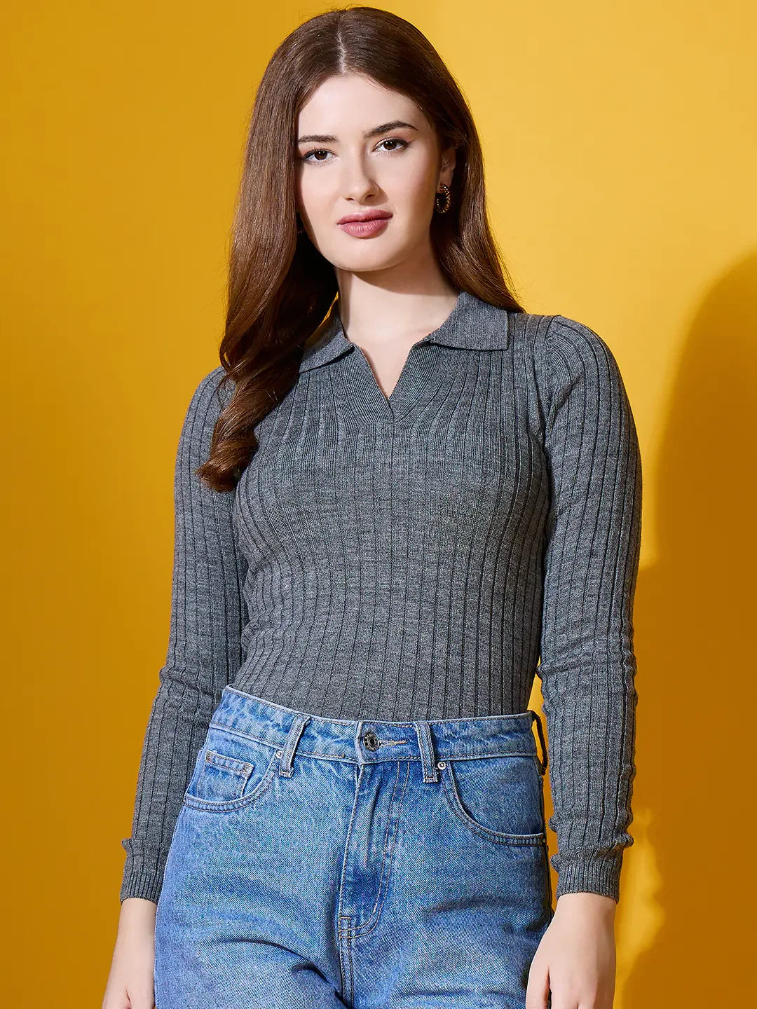 Woman with long brown hair wears 98degreenorth’s grey ribbed collar sweater with blue jeans against a yellow background.