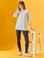 A woman stands against a yellow background, wearing a light tunic and 98degreenorth Women Navy Ankle-Length Leggings. She touches her hair with one hand and rests the other on a tall white stool, completing the look with sandals.