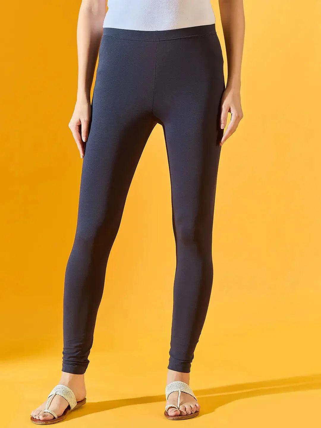 A person wearing 98degreenorth Women Navy Ankle-Length Leggings, paired with a white top and beige sandals, stands against a solid yellow background. The image is cropped at the shoulders.