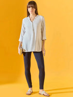 A woman stands against a yellow background, wearing a light-colored long-sleeve tunic, 98degreenorth Women Navy Ankle-Length Leggings with stretchable fit, sandals, and holding a small clutch.