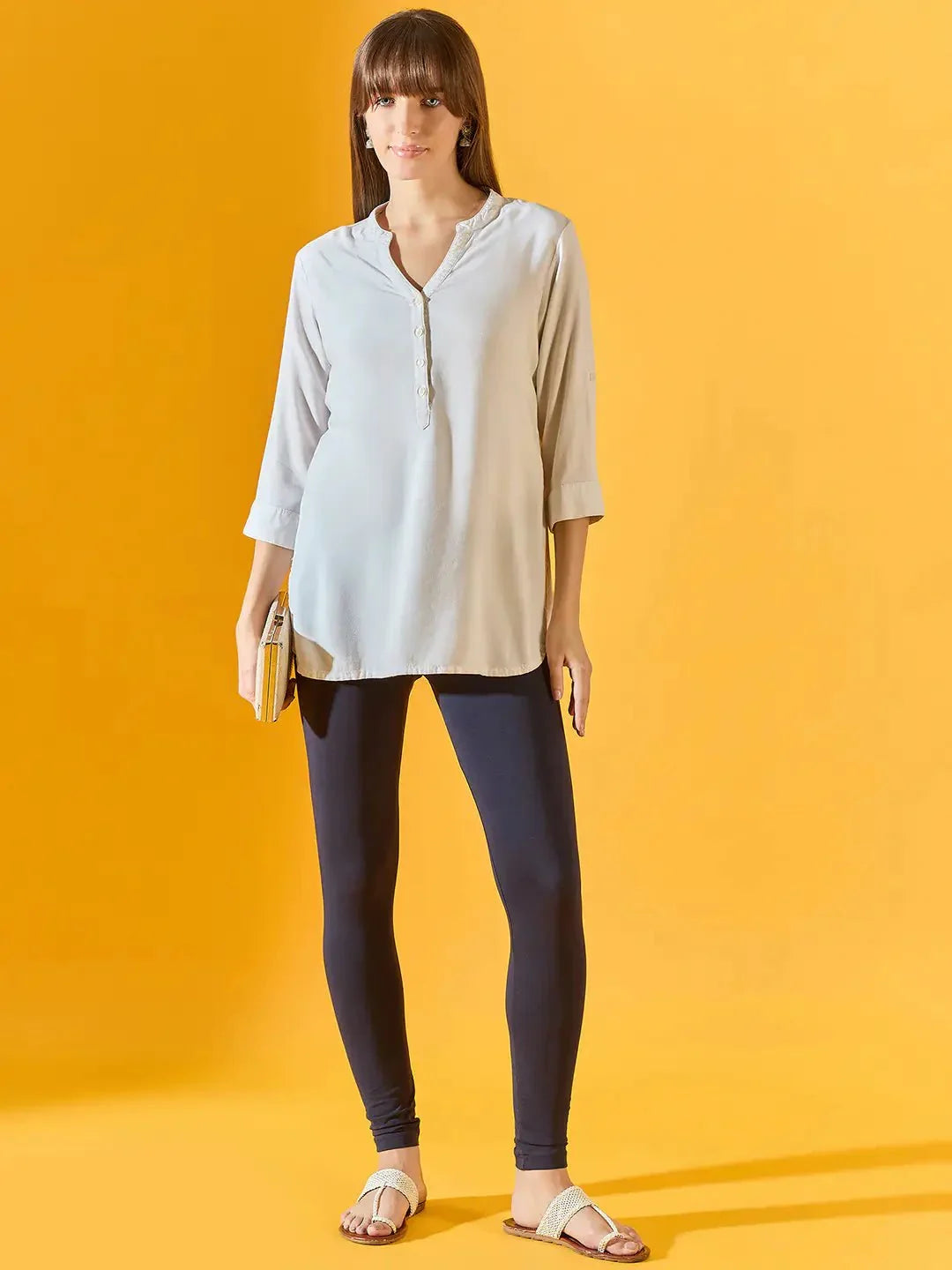 A woman stands against a yellow background, wearing a light-colored long-sleeve tunic, 98degreenorth Women Navy Ankle-Length Leggings with stretchable fit, sandals, and holding a small clutch.