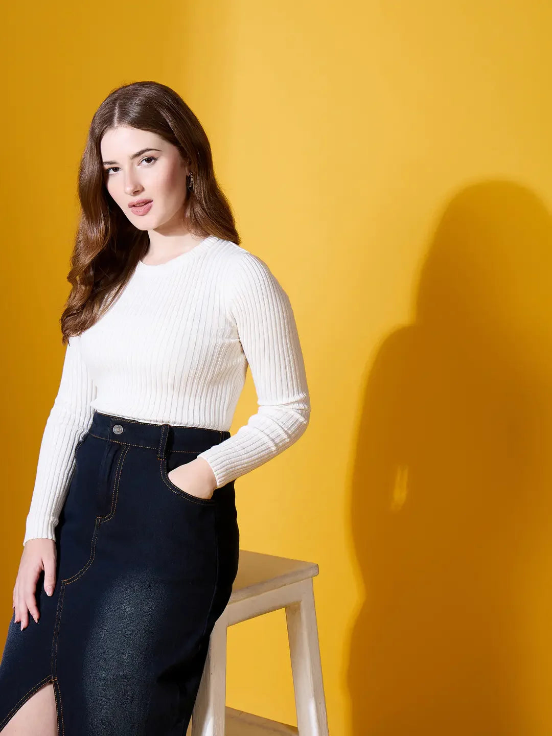Woman models 98degreenorth Off White Round Neck Ribbed Sweater by a stool against a bright yellow background.