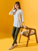 Woman in white tunic and 98degreenorth Women Olive Ankle-Length Leggings stands by wooden chair against yellow background.