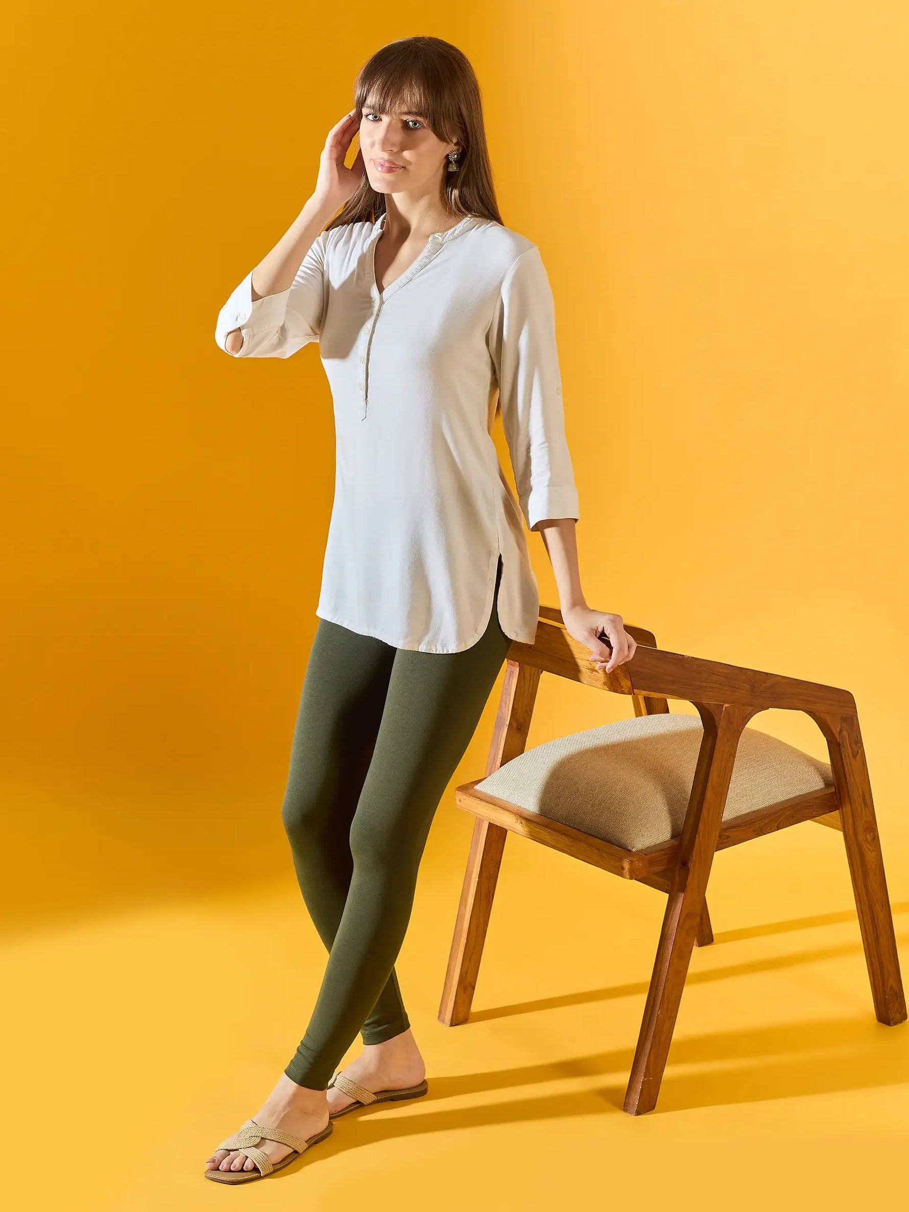 Woman in white tunic and 98degreenorth Women Olive Ankle-Length Leggings stands by wooden chair against yellow background.