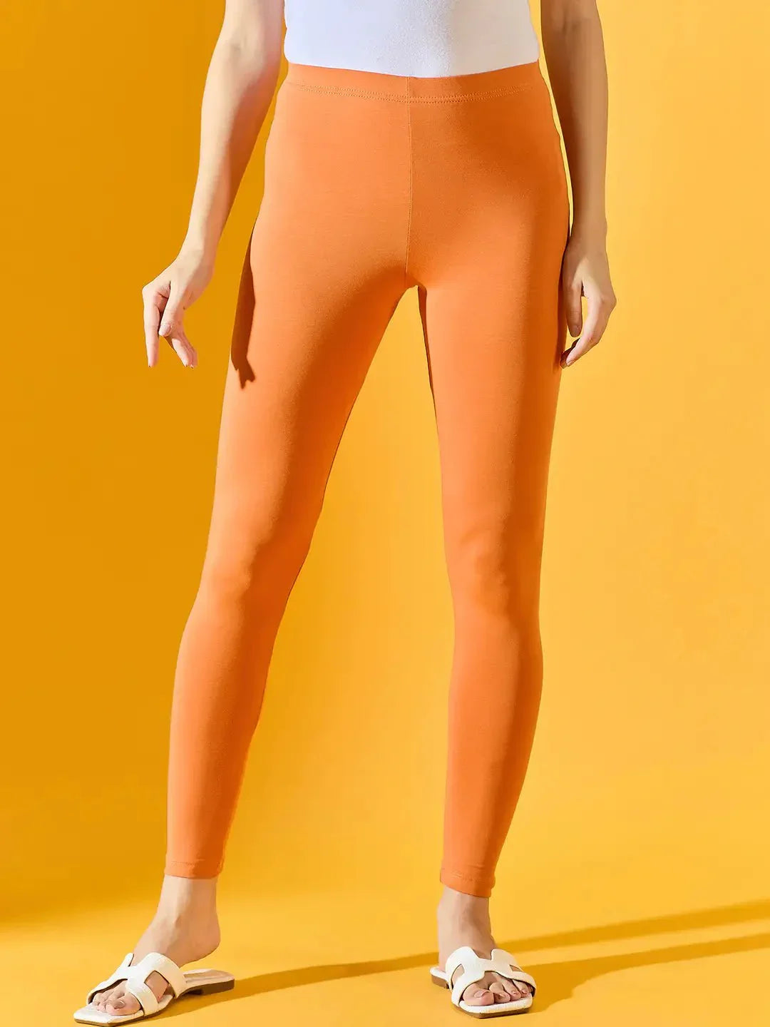 A person wears 98degreenorth Women Orange Ankle-Length Leggings, a white top, and sandals against a yellow background.