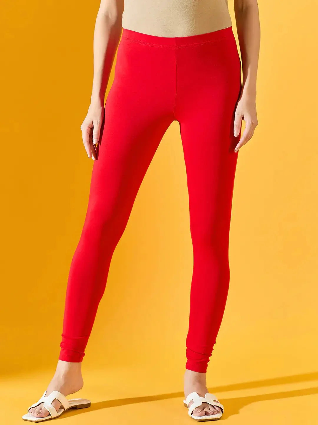 Only the lower half of a person is visible, wearing 98degreenorth Women Red Ankle-Length Leggings against a yellow background, paired with a beige top and white slide sandals.