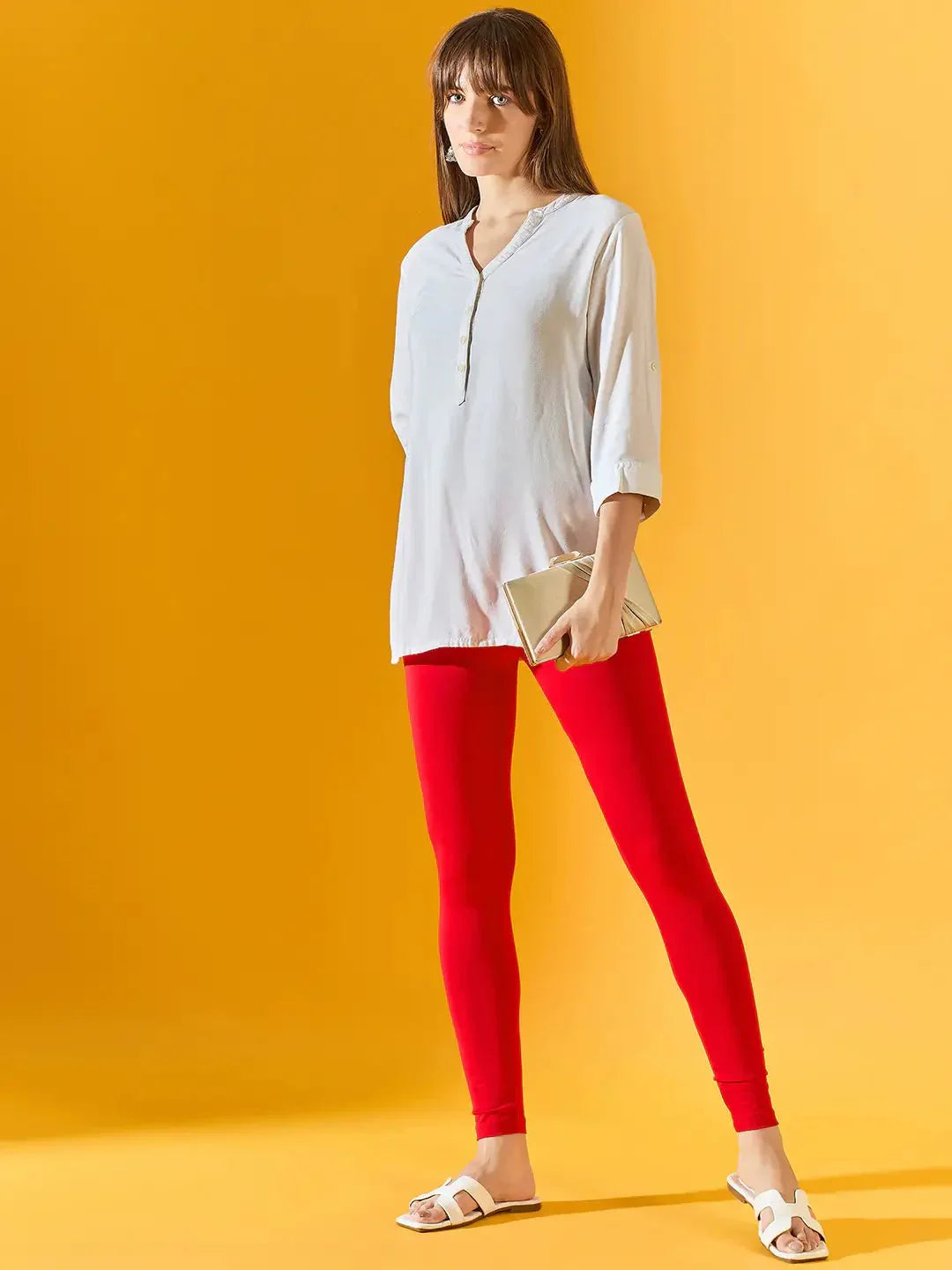 A woman stands against a yellow background wearing 98degreenorth Women Red Ankle-Length Leggings, a loose white tunic, white sandals, and holding a beige clutch.