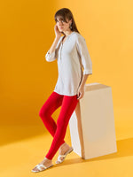 A woman in 98degreenorth’s Women Red Ankle-Length Leggings stands against a yellow background, smiling and leaning on a white block; she wears a light gray tunic and white sandals.