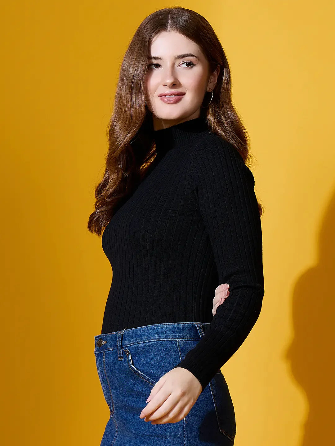 Woman with long brown hair in 98degreenorth’s black ribbed high neck sweater stands smiling against a yellow background.