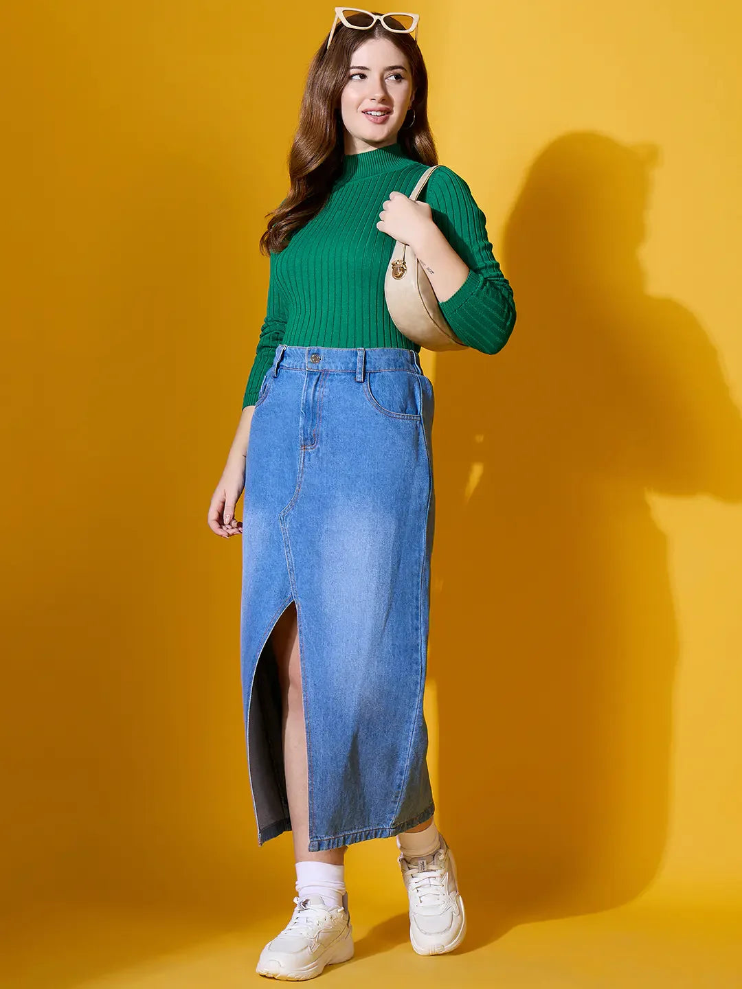 Woman in a 98degreenorth emerald green ribbed high neck sweater, denim skirt, and white sneakers stands against yellow.
