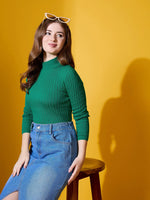A woman in a 98degreenorth emerald green ribbed high neck sweater and denim skirt sits on a stool, yellow background.