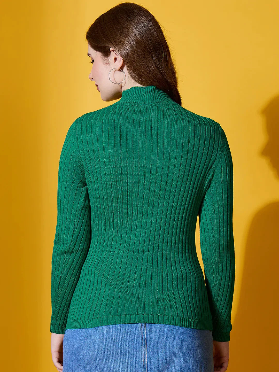 Woman wears 98degreenorth Ribbed High Neck Sweater in emerald green, standing with her back to a yellow background.